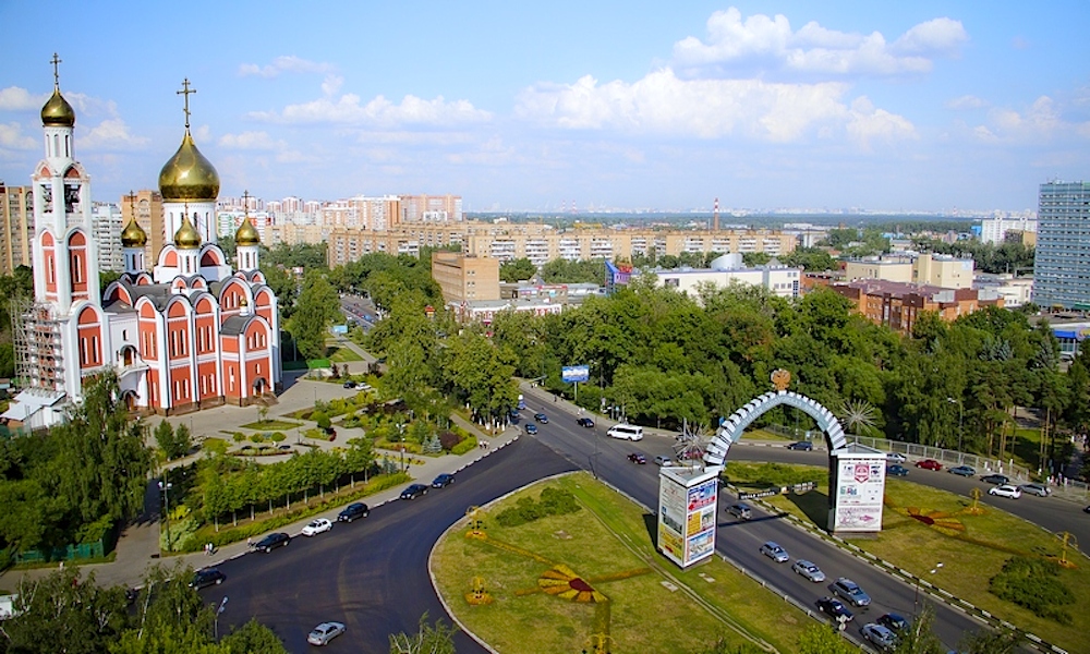 На фотографии изображен городской пейзаж. Слева находится крупная церковь в традиционном русском православном стиле. У церкви несколько золотых куполов и стены красного и белого цветов. Перед церковью расположен ухоженный сад с дорожками и зелеными насаждениями. Справа от церкви находится оживленное кольцевое движение, по которому едут несколько автомобилей. На кольце установлена декоративная арка с рекламными щитами по обеим сторонам. За кольцевым движением видны деревья и здания, среди которых есть как жилые, так и коммерческие постройки. На заднем плане простирается городской район с множеством многоквартирных домов. Небо ясное, с небольшим количеством облаков.