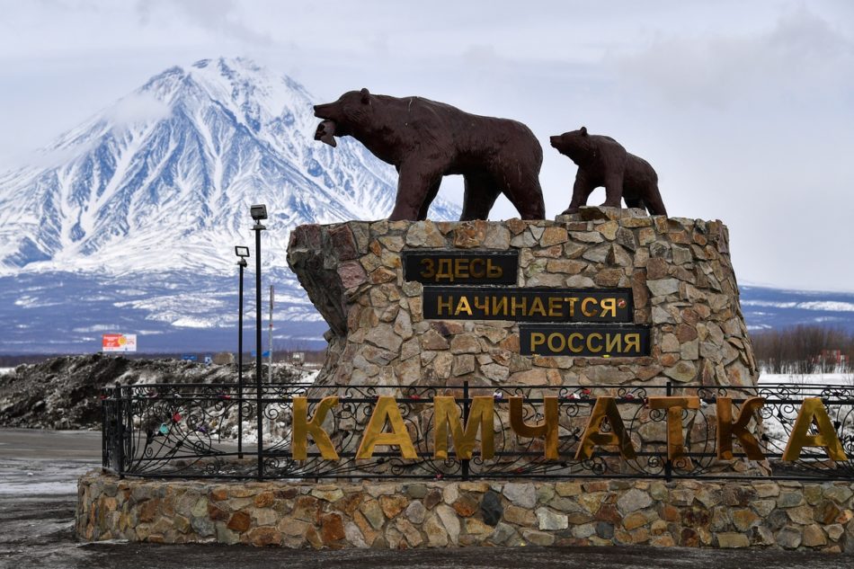 На изображении показан памятник, расположенный на Камчатке. Памятник представляет собой скульптуру двух медведей, стоящих на каменной платформе. На платформе написано: "Здесь начинается Россия". На переднем плане также видна надпись "Камчатка". На заднем плане виден заснеженный вулкан или гора, что подчеркивает природную красоту региона.