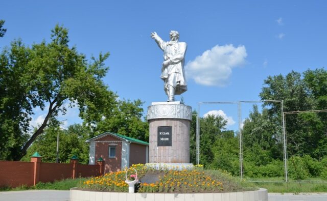 На фотографии изображен памятник Кузьме Минину, расположенный на открытой площадке. Памятник состоит из статуи и постамента. Статуя выполнена из металла и изображает Кузьму Минина в полный рост. Он одет в традиционную одежду того времени: длинный кафтан с поясом и сапоги. Правая рука Минина поднята и указывает вперёд, как будто он призывает к действию. Левая рука прижата к груди, что может символизировать преданность и решимость. Статуя стоит на высоком цилиндрическом постаменте, который украшен барельефами. На передней части постамента есть чёрная табличка с белой надписью "Кузьма Минин". Вокруг постамента высажены цветы, которые образуют круглый цветник. Цветы яркие, преимущественно жёлтые и оранжевые, что добавляет красок к общей картине. Небо на фотографии ясное, с несколькими белыми облаками, что создаёт ощущение солнечного и тёплого дня.