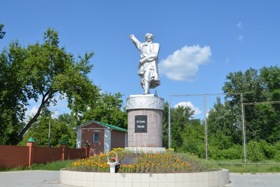 На фотографии изображен памятник Кузьме Минину, расположенный на открытой площадке. Памятник состоит из статуи и постамента. Статуя выполнена из металла и изображает Кузьму Минина в полный рост. Он одет в традиционную одежду того времени: длинный кафтан с поясом и сапоги. Правая рука Минина поднята и указывает вперёд, как будто он призывает к действию. Левая рука прижата к груди, что может символизировать преданность и решимость. Статуя стоит на высоком цилиндрическом постаменте, который украшен барельефами. На передней части постамента есть чёрная табличка с белой надписью "Кузьма Минин". Вокруг постамента высажены цветы, которые образуют круглый цветник. Цветы яркие, преимущественно жёлтые и оранжевые, что добавляет красок к общей картине. Небо на фотографии ясное, с несколькими белыми облаками, что создаёт ощущение солнечного и тёплого дня.