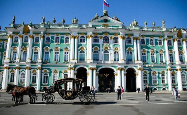 На фотографии изображен Зимний дворец в Санкт-Петербурге, Россия. Это здание имеет три этажа и выполнено в стиле барокко. Стены дворца окрашены в бирюзовый цвет, а окна и колонны выделены белым. Колонны украшены золотыми элементами, которые придают зданию роскошный вид. На крыше дворца расположены статуи, изображающие различных мифологических персонажей. В центре здания находится главный вход, который представлен тремя большими арками. Над центральной аркой виден российский флаг, развевающийся на ветру. Также над арками можно увидеть часы. Перед дворцом на площади стоит черная карета с золотыми украшениями, запряженная двумя лошадьми. Карета имеет классический дизайн с большими колесами и гербом на дверях. Лошади стоят спокойно, а рядом с ними находится человек, вероятно, кучер. На площади перед дворцом ходят люди, некоторые из них фотографируются, другие просто прогуливаются. Погода ясная, небо чистое и голубое, что подчеркивает яркость и красоту здания.