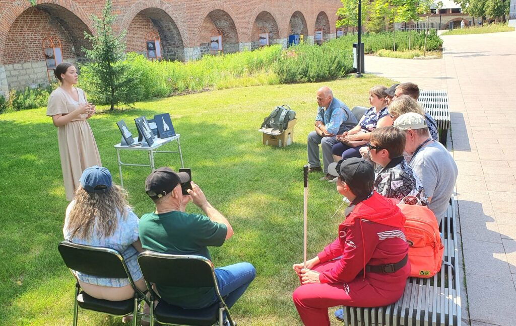 На фотографии изображена сцена на открытом воздухе в Нижегородском кремле. На переднем плане, слева направо, сидят несколько человек. Некоторые из них держат белые трости. Перед этой группой стоит женщина в светлом длинном платье. Она что-то рассказывает и жестикулирует руками. Перед ней на небольшом складном столике расположены несколько рельефных макетов, которые, предназначены для тактильного ознакомления. 