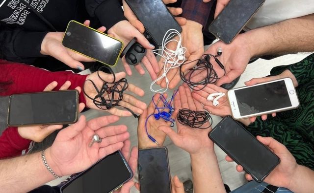 Группа людей стоит в круге, вытянув руки к центру. В руках у каждого мобильный телефон и наушники.