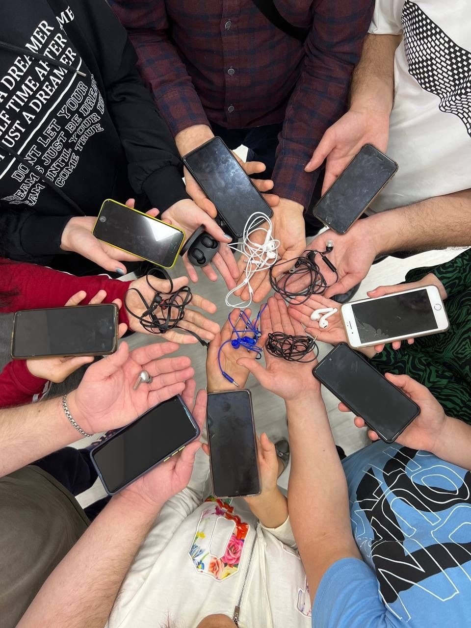 Группа людей стоит в круге, вытянув руки к центру. В руках у каждого мобильный телефон и наушники.