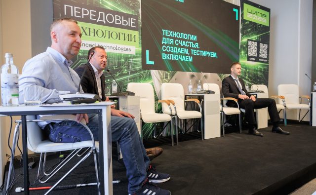 Три мужчины на сцене во время деловой панели. Слева мужчина среднего возраста, светлая кожа, короткие светлые волосы, в голубой рубашке и синих джинсах, в тёмно-синих кроссовках Adidas с белыми полосками. Он сидит за белым столом на белом стуле с металлическими ножками; на столе прозрачный пустой стакан, пластиковая бутылка воды и настольный микрофон/пульт конференц-связи. На полу у его ног лежит белая трость с красным концом. Рядом, чуть правее, второй мужчина, тоже светлокожий, в тёмном пиджаке и светлой рубашке без галстука, сидит в белом кресле; перед ним белая тумба с микрофоном и бутылкой воды. Справа на сцене третий мужчина, светлая кожа, короткие светлые волосы, он в тёмном костюме, белой рубашке и галстуке, сидит в белом кресле с деревянными подлокотниками; рядом такая же тумба с микрофоном и бутылкой воды. На сцене ещё несколько пустых одинаковых белых кресел. За спинами участников крупный баннер в зелёно-чёрных тонах с абстрактными линиями, напоминающими электронные схемы. Слева вертикальный баннер с надписью крупным белым шрифтом: «ПЕРЕДОВЫЕ ТЕХНОЛОГИИ» Под ним текст меньшим шрифтом на английском: «Advanced technologies». В центре над сценой большой экран с чёрным фоном и зелёными линиями, на котором светло-зелёными буквами написано: «ТЕХНОЛОГИИ ДЛЯ СЧАСТЬЯ. СОЗДАЁМ, ТЕСТИРУЕМ, ВКЛЮЧАЕМ».