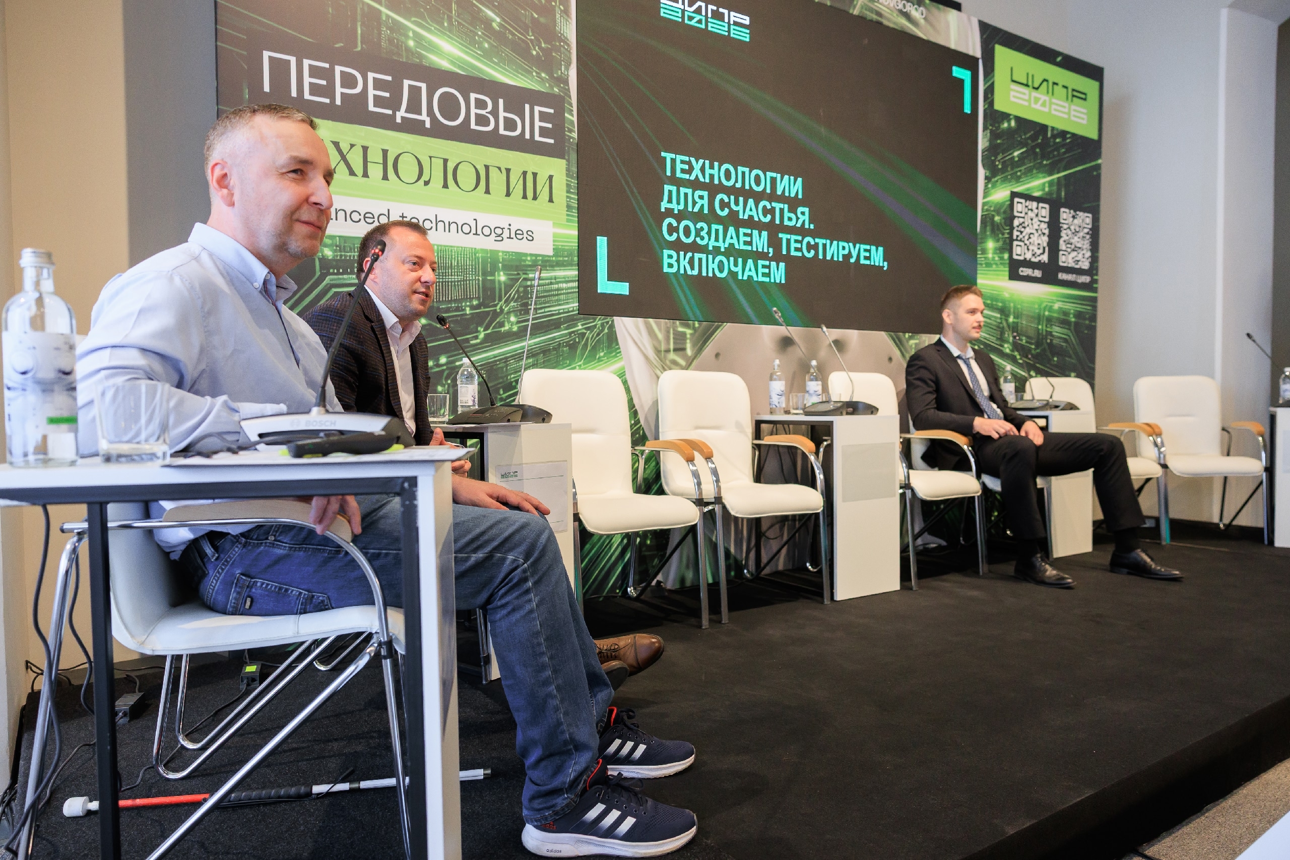 Три мужчины на сцене во время деловой панели. Слева мужчина среднего возраста, светлая кожа, короткие светлые волосы, в голубой рубашке и синих джинсах, в тёмно-синих кроссовках Adidas с белыми полосками. Он сидит за белым столом на белом стуле с металлическими ножками; на столе прозрачный пустой стакан, пластиковая бутылка воды и настольный микрофон/пульт конференц-связи. На полу у его ног лежит белая трость с красным концом. Рядом, чуть правее, второй мужчина, тоже светлокожий, в тёмном пиджаке и светлой рубашке без галстука, сидит в белом кресле; перед ним белая тумба с микрофоном и бутылкой воды. Справа на сцене третий мужчина, светлая кожа, короткие светлые волосы, он в тёмном костюме, белой рубашке и галстуке, сидит в белом кресле с деревянными подлокотниками; рядом такая же тумба с микрофоном и бутылкой воды. На сцене ещё несколько пустых одинаковых белых кресел. За спинами участников крупный баннер в зелёно-чёрных тонах с абстрактными линиями, напоминающими электронные схемы. Слева вертикальный баннер с надписью крупным белым шрифтом: «ПЕРЕДОВЫЕ ТЕХНОЛОГИИ» Под ним текст меньшим шрифтом на английском: «Advanced technologies». В центре над сценой большой экран с чёрным фоном и зелёными линиями, на котором светло-зелёными буквами написано: «ТЕХНОЛОГИИ ДЛЯ СЧАСТЬЯ. СОЗДАЁМ, ТЕСТИРУЕМ, ВКЛЮЧАЕМ».