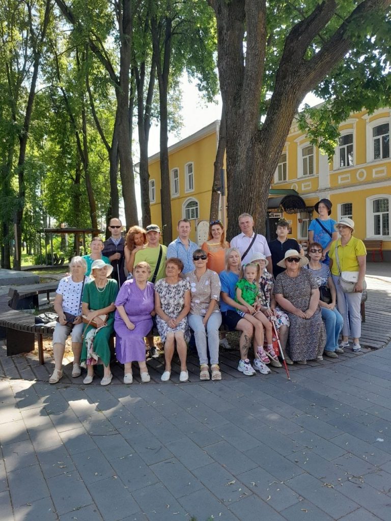 Групповое фото на улице. На снимке около двадцати человек разного возраста, большинство из них — женщины, есть несколько мужчин и дети. Люди сидят и стоят на скамейке под большими деревьями, на заднем плане виден желтый двухэтажный дом с белыми окнами и декоративными элементами. День солнечный, вокруг много зелени, тени от деревьев. Все выглядят расслабленными и улыбаются, создаётся впечатление дружеской встречи или экскурсии.