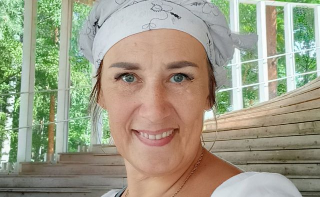 На фото женщина среднего возраста с голубыми глазами, светлой кожей и светло-коричневыми волосами, частично покрытыми белым платком с чёрным цветочным орнаментом. Она улыбается и одета в белую блузу, на шее у неё цепочка. На заднем плане — деревянные ступени амфитеатра или павильона с большими окнами, за которыми виднеется зелёный лес, солнечно и светло.