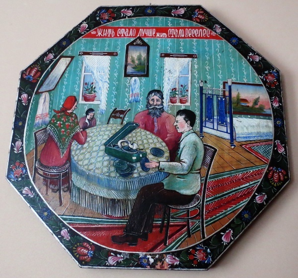 Панно в стиле городецкой росписи. Изображена семья из четырёх человек, сидящих за круглым столом, покрытым скатертью. На столе стоит патефон, и один из мужчин заводит его. В комнате зелёные обои с белым узором, два окна с кружевными занавесками, на подоконниках стоят цветы. На стене между окнами висит картина. На полу лежит яркий ковёр, справа видна кровать с покрывалом и ещё одна картина на стене. В верхней части шкатулки написано: "Жить стало лучше, жить стало веселее". Края украшены цветочным орнаментом.