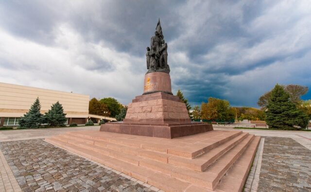 На фотографии изображен памятник "Молодогвардейцам" в городе Краснодон. Памятник представляет собой скульптурную композицию, установленную на массивном постаменте из красного камня. Скульптура изображает несколько фигур молодых людей, стоящих вместе, символизируя единство и героизм членов организации "Молодая гвардия". Фигуры выполнены в реалистичном стиле, и каждая из них выражает решимость и мужество. Постамент, на котором установлена скульптура, имеет несколько ступеней, ведущих к основанию памятника. Вокруг памятника выложена брусчатка, создавая просторную площадь для посетителей. На заднем плане видны деревья и здание светлого цвета, что подчеркивает монументальность и значимость памятника. Небо на фотографии затянуто облаками, что добавляет драматичности общей атмосфере. На переднем плане видны ступени, ведущие к памятнику. Они выполнены из того же красного камня, что и постамент. Площадка вокруг памятника выложена брусчаткой, создавая контраст с гладкими ступенями. Справа и слева от памятника растут зеленые деревья, добавляя природный элемент в композицию. Здание на заднем плане имеет светлый фасад и современный архитектурный стиль. Оно расположено слева от памятника и частично скрыто деревьями. Небо над памятником затянуто плотными серыми облаками, создавая ощущение надвигающейся грозы или дождя. Это придает фотографии драматичное и торжественное настроение.