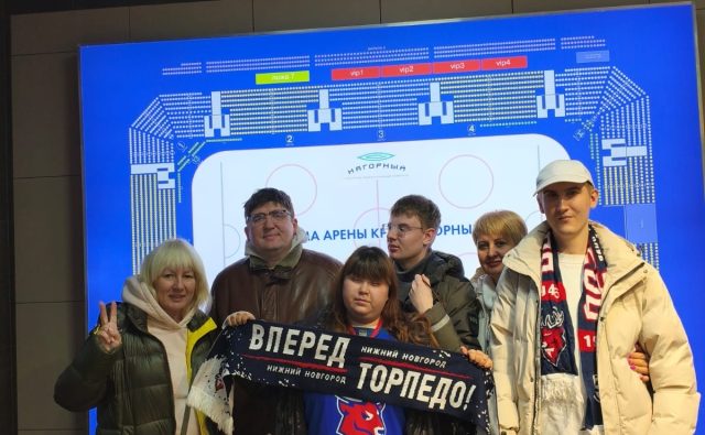 Группа из шести человек стоит перед большим электронным табло с планом хоккейной арены “Нагорный”. На переднем плане девочка держит шарф с надписью «ВПЕРЕД ТОРПЕДО! НИЖНИЙ НОВГОРОД».