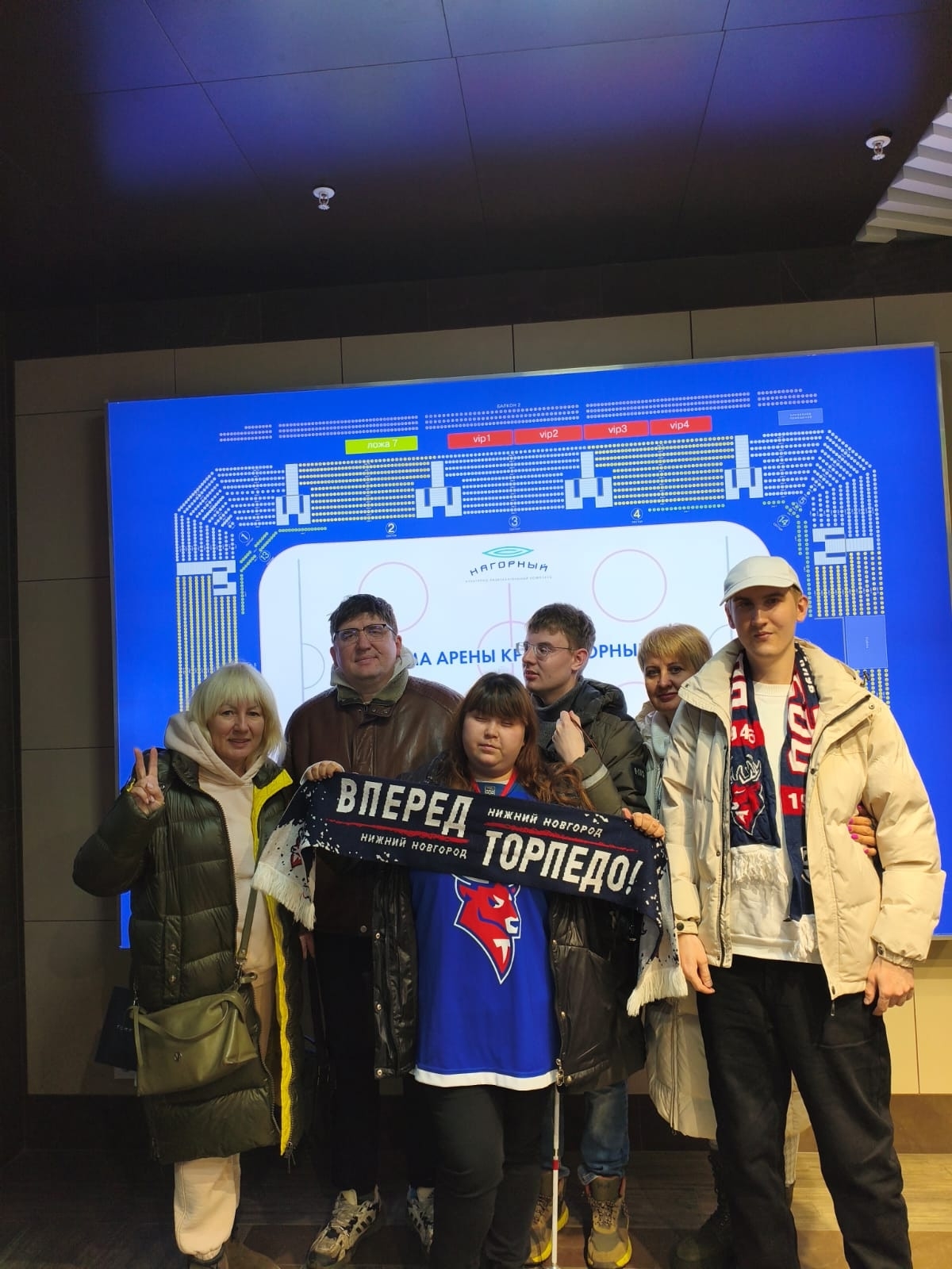 Группа из шести человек стоит перед большим электронным табло с планом хоккейной арены “Нагорный”. На переднем плане девочка держит шарф с надписью «ВПЕРЕД ТОРПЕДО! НИЖНИЙ НОВГОРОД».