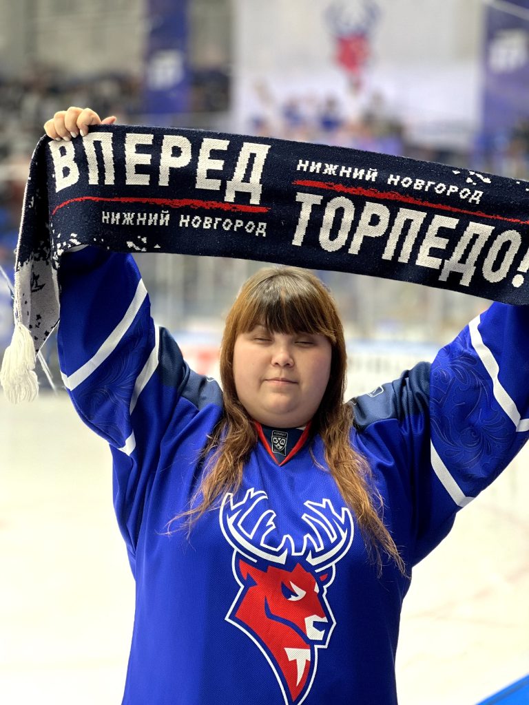Болельщица в синем хоккейном свитере с изображением красного оленя держит шарф над головой. На шарфе белыми буквами написано: "Вперёд Торпедо! Нижний Новгород".