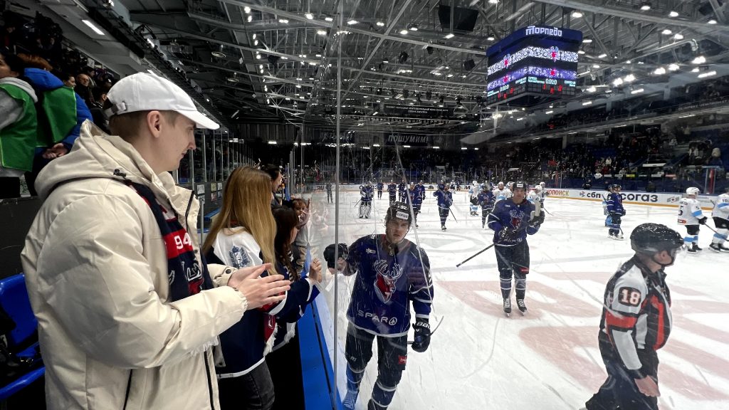 В центре изображения — хоккеист в синей форме с оленьей эмблемой (клуб Торпедо), который приветствует зрителей
через стекло.