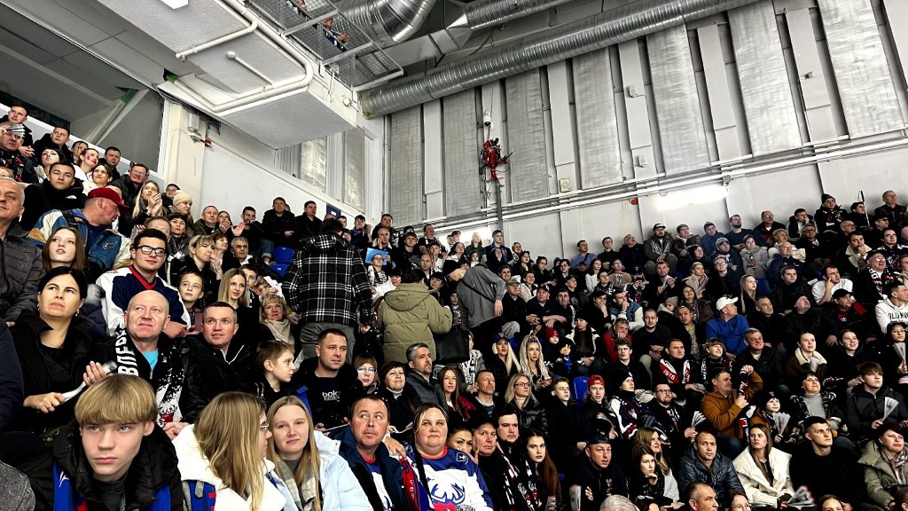 Трибуна ледового дворца, заполненная болельщиками. Люди разного возраста сидят плотно друг к другу, многие одеты в тёплую одежду и шарфы с символикой команды,
некоторые держат бело-синие и черные шарфы с надписями. На лицах выражения ожидания, интереса, кто-то улыбается, кто-то смотрит вперёд с сосредоточенностью.