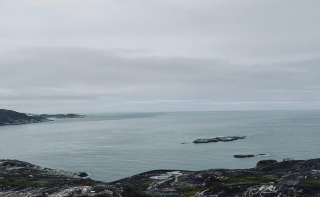 На фотографии изображен морской пейзаж. На переднем плане видны скалистые берега с редкой растительностью. В центре фотографии простирается спокойное море, а на заднем плане виднеется береговая линия с холмами и скалами. Небо облачное и серое, создавая атмосферу прохладного и пасмурного дня.