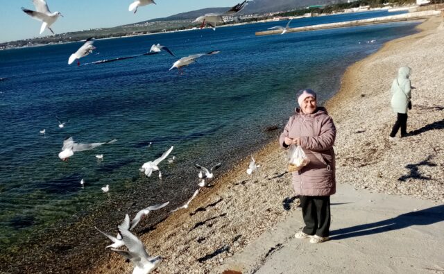 На фотографии изображен морской берег с галькой и бетонной набережной. В центре кадра стоит женщина, одетая в розовую куртку и розовую шапку. Она держит в руках прозрачный пакет, в котором, вероятно, находится корм для птиц. Женщина улыбается и смотрит в сторону камеры. Вокруг женщины летают чайки. Некоторые из них парят в воздухе с расправленными крыльями, другие сидят на гальке или на бетонной набережной. Чайки белые с черными кончиками крыльев и красными клювами. На переднем плане, ближе к нижнему краю фотографии, стоит голубь, который смотрит в сторону камеры. На заднем плане, ближе к правому краю фотографии, стоит еще один человек. Этот человек одет в светло-зеленую куртку и светлую шапку. Он стоит спиной к камере и смотрит на море. Море спокойное, его воды темно-синие и прозрачные. Вода простирается до горизонта, где видны горы и здания. Горы покрыты зеленью, а здания расположены вдоль берега. Небо ясное и голубое, без облаков, что указывает на солнечную погоду. На переднем плане, ближе к нижнему краю фотографии, видна бетонная набережная, которая переходит в гальку. Галька мелкая и светлая, покрывает весь берег. Тени от людей и птиц падают на бетонную набережную, что указывает на яркое солнечное освещение.