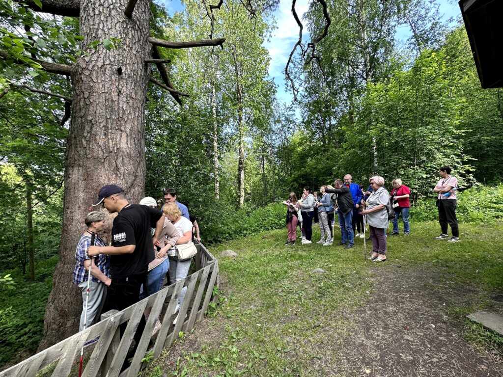 На фотографии изображена группа людей, находящихся на природе. Они стоят на травянистой поляне, окруженной деревьями и кустарниками. В центре внимания — большое дерево с толстыми ветвями и корой, к которому прислонились несколько человек. Некоторые из них держат белые трости, что может указывать на то, что они незрячие или слабовидящие. Люди одеты в повседневную одежду, и кажется, что они участвуют в каком-то мероприятии или экскурсии. На переднем плане виден деревянный заборчик, за которым стоят несколько человек. Погода ясная, небо голубое с небольшим количеством облаков.