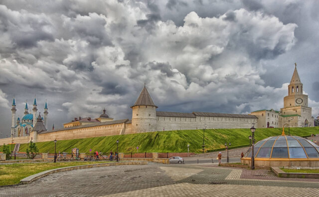 На фотографии изображен Казанский Кремль в Казани, Россия. На переднем плане видна часть площади с мощеными дорожками и стеклянным куполом, который, вероятно, является входом в подземное помещение. В центре фотографии находится крепостная стена с башнями, а на заднем плане слева виднеется мечеть Кул-Шариф с голубыми куполами и минаретами. Справа видна Спасская башня с часами. Небо покрыто плотными серыми облаками, создавая драматичную атмосферу. На площади можно увидеть несколько людей, гуляющих и катающихся на велосипедах.