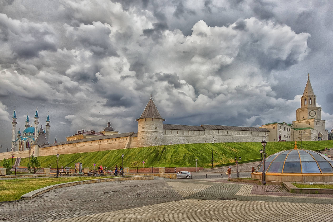 На фотографии изображен Казанский Кремль в Казани, Россия. На переднем плане видна часть площади с мощеными дорожками и стеклянным куполом, который, вероятно, является входом в подземное помещение. В центре фотографии находится крепостная стена с башнями, а на заднем плане слева виднеется мечеть Кул-Шариф с голубыми куполами и минаретами. Справа видна Спасская башня с часами. Небо покрыто плотными серыми облаками, создавая драматичную атмосферу. На площади можно увидеть несколько людей, гуляющих и катающихся на велосипедах.