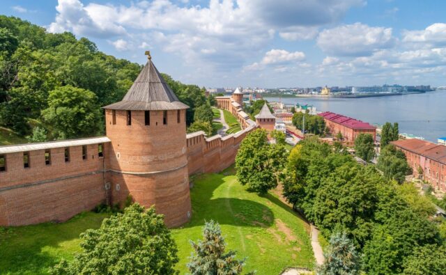 На фотографии изображен панорамный вид на Нижегородский кремль и прилегающие к нему территории. На переднем плане, слева, находится массивная круглая башня кремля, построенная из красного кирпича. Башня имеет конусообразную крышу, покрытую серыми деревянными досками. На вершине крыши установлен флюгер в виде креста. Башня соединена с длинной кирпичной стеной, которая тянется вдоль холма и уходит вдаль, постепенно уменьшаясь в перспективе. В стене расположены узкие вертикальные бойницы. Справа от башни и стены находится зеленая территория с густыми деревьями и кустарниками. Внизу видна тропинка, ведущая вдоль стены кремля. Далее, за стеной кремля, видны несколько других башен, также построенных из красного кирпича и с конусообразными крышами. Эти башни расположены на разном расстоянии друг от друга и соединены стеной. На заднем плане, справа, виден город с разноцветными зданиями. Некоторые здания имеют красные крыши, другие – белые или зеленые. Среди них выделяется белое здание с золотым куполом, вероятно, церковь или собор. Справа от города протекает широкая река Волга. На противоположном берегу реки видны современные здания и сооружения. Небо ясное, голубое, с небольшими пушистыми облаками, создающими приятный контраст с зеленью деревьев и красным кирпичом кремля.