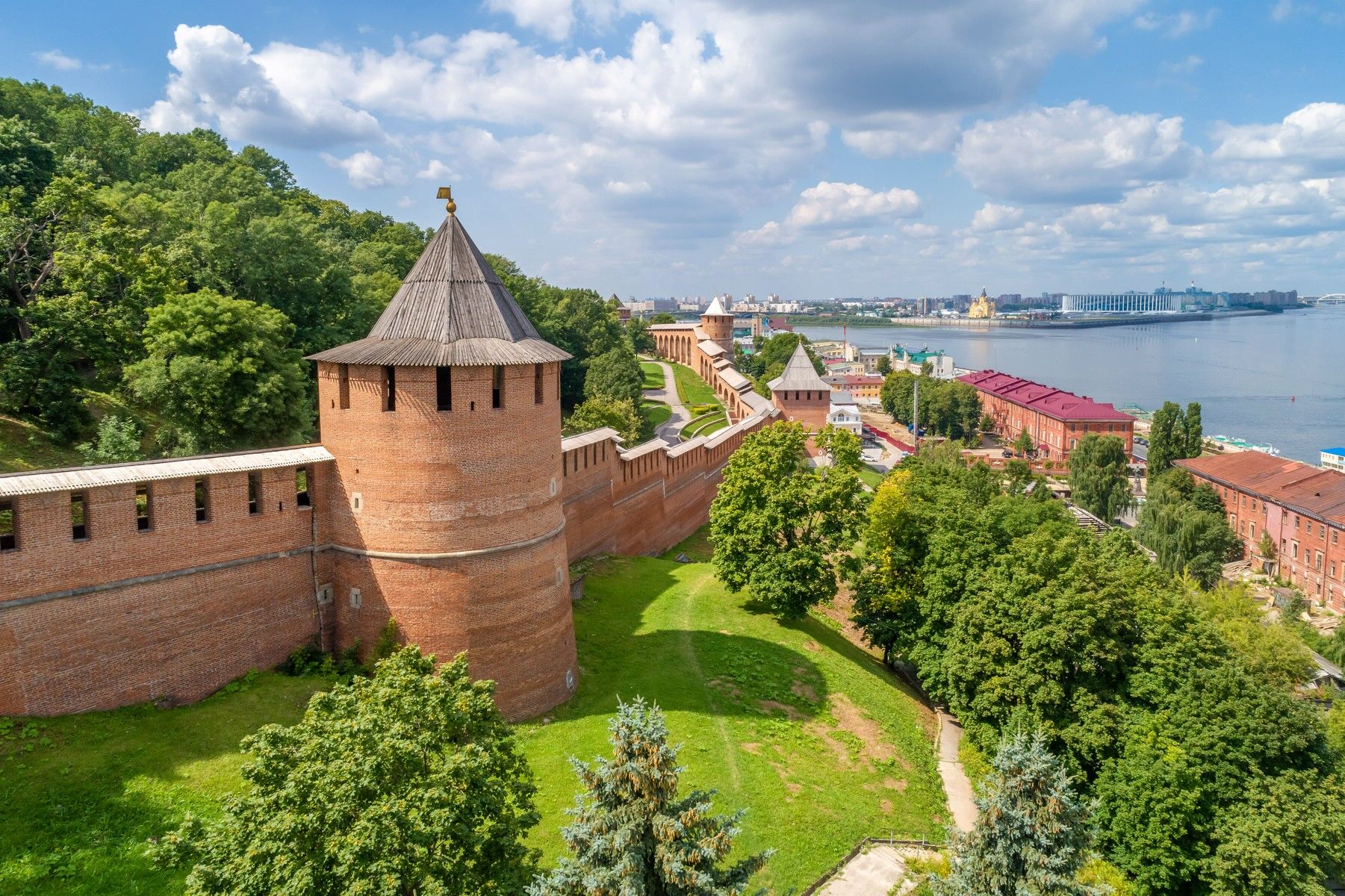 На фотографии изображен панорамный вид на Нижегородский кремль и прилегающие к нему территории. На переднем плане, слева, находится массивная круглая башня кремля, построенная из красного кирпича. Башня имеет конусообразную крышу, покрытую серыми деревянными досками. На вершине крыши установлен флюгер в виде креста. Башня соединена с длинной кирпичной стеной, которая тянется вдоль холма и уходит вдаль, постепенно уменьшаясь в перспективе. В стене расположены узкие вертикальные бойницы. Справа от башни и стены находится зеленая территория с густыми деревьями и кустарниками. Внизу видна тропинка, ведущая вдоль стены кремля. Далее, за стеной кремля, видны несколько других башен, также построенных из красного кирпича и с конусообразными крышами. Эти башни расположены на разном расстоянии друг от друга и соединены стеной. На заднем плане, справа, виден город с разноцветными зданиями. Некоторые здания имеют красные крыши, другие – белые или зеленые. Среди них выделяется белое здание с золотым куполом, вероятно, церковь или собор. Справа от города протекает широкая река Волга. На противоположном берегу реки видны современные здания и сооружения. Небо ясное, голубое, с небольшими пушистыми облаками, создающими приятный контраст с зеленью деревьев и красным кирпичом кремля.