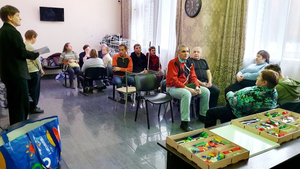 Комната, в которой находятся люди, сидящие на стульях в несколько небольших групп. Некоторые люди одеты в повседневную одежду, кто-то держит трость для
незрячих. Справа у окна мужчина и женщина беседуют. На переднем плане стол с коробками, наполненными цветными геометрическими фигурами из пластика или
дерева. В левой части кадра стоят двое человек, один из них держит папку или тетрадь. На стене часы и телевизор, в углу видна проволочная фигура, похожая
на оленя. Обстановка напоминает занятие или собрание в центре социального обслуживания или реабилитации для людей с нарушением зрения.