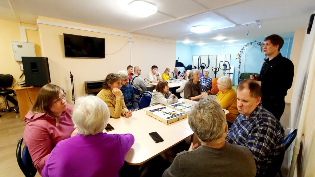 Комната с несколькими столами, за которыми сидят взрослые люди и пожилые, около 18 человек. Большинство сидят, некоторые стоят, все вовлечены в беседу
или слушают мужчину, который стоит справа и что-то объясняет или рассказывает. На столах лежат магнитно-тактильные конструкторы или рельефно-графические схемы.