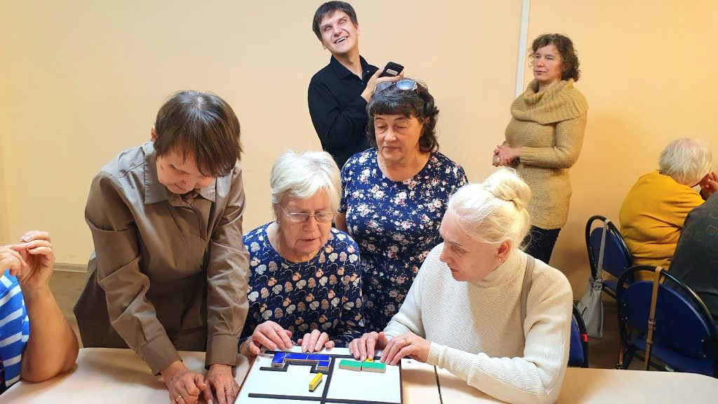 Группа людей за столом. На переднем плане четыре женщины, которые увлечённо трогают пальцами тактильную схему с разноцветными плоскими фигурами на белой
поверхности. Справа от них сидит женщина в светлом свитере с белыми волосами, собранными в пучок. Слева – женщина с короткой стрижкой наклоняется вперёд,
помогая. За столом стоят ещё две пожилые женщины, одна из них в синем платье с цветочным принтом. На заднем плане мужчина в чёрной рубашке улыбается и
держит телефон. Рядом с ним стоит женщина в бежевом свитере с высоким воротником и улыбается. В правой части кадра, за другим столом, видны ещё две женщины
– одна из них в жёлтом свитере. Обстановка напоминает уютную комнату для совместного досуга или занятий.