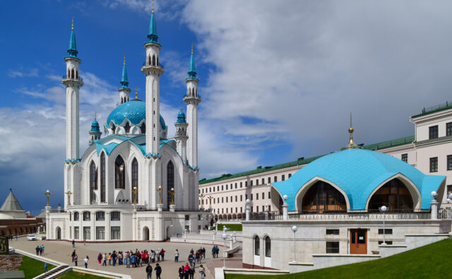 На фотографии изображена мечеть Кул-Шариф, расположенная в Казанском Кремле, Татарстан, Россия. Мечеть имеет четыре высоких минарета с голубыми куполами и белыми стенами. Справа от мечети находится здание с голубой крышей, которое также является частью комплекса. На переднем плане видна группа людей, гуляющих по площади перед мечетью. Небо частично облачное, с проблесками синего цвета.