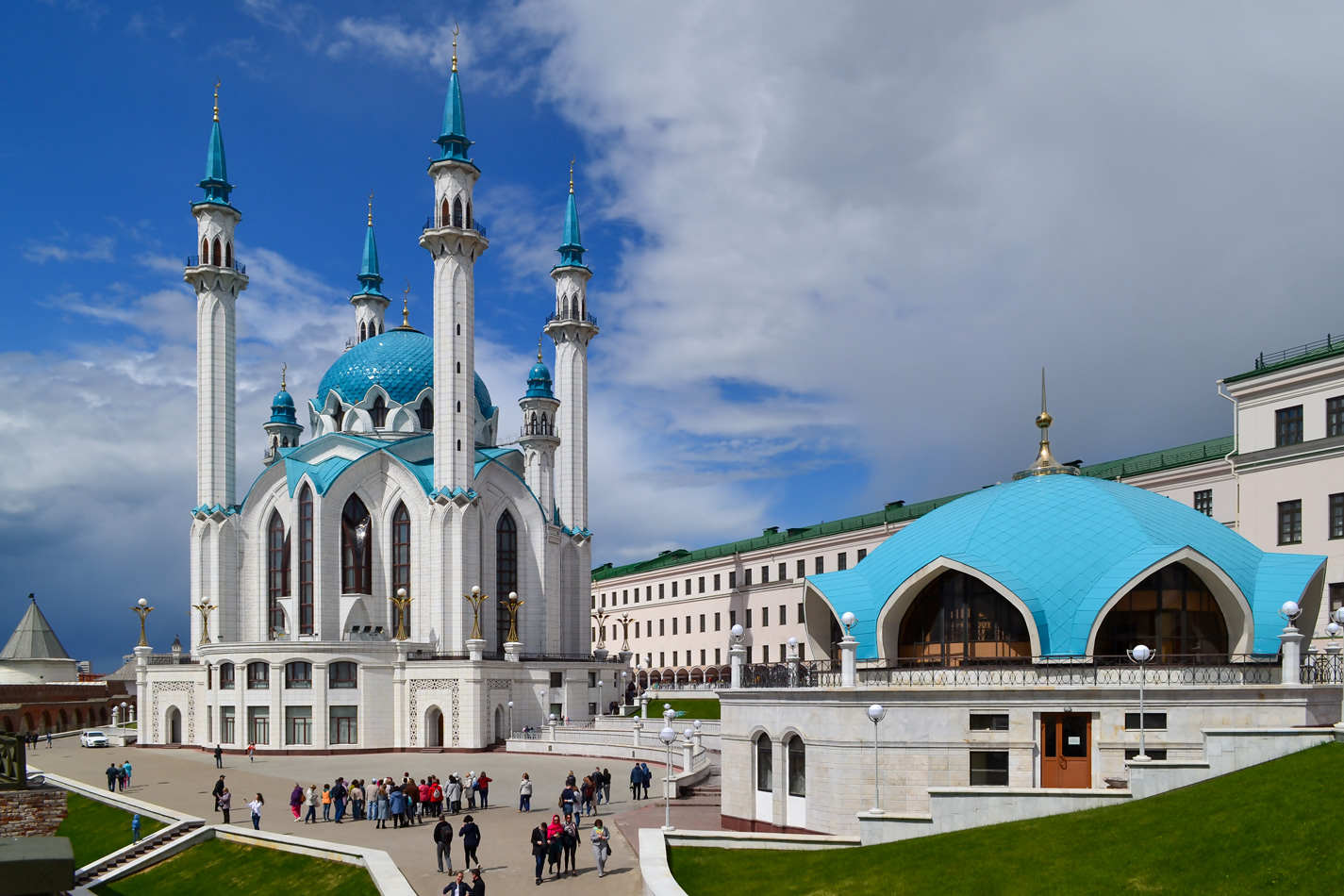 На фотографии изображена мечеть Кул-Шариф, расположенная в Казанском Кремле, Татарстан, Россия. Мечеть имеет четыре высоких минарета с голубыми куполами и белыми стенами. Справа от мечети находится здание с голубой крышей, которое также является частью комплекса. На переднем плане видна группа людей, гуляющих по площади перед мечетью. Небо частично облачное, с проблесками синего цвета.