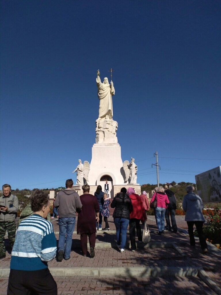 На фотографии изображена группа из примерно двадцати человек, стоящих на улице в парке. Они собрались на каменной дорожке возле невысокой каменной стены, за которой находится водоем. Водоем окружен камнями, и вода в нем спокойная. Люди одеты в осеннюю одежду: куртки, свитера, шарфы. Некоторые из них держат в руках фотоаппараты, сумки и телефоны. В центре группы стоит женщина, вероятно, экскурсовод, которая что-то рассказывает. Она одета в темную юбку и светлый жакет. Люди вокруг нее внимательно слушают, некоторые смотрят в ее сторону, другие делают фотографии. На заднем плане видны деревья с зелеными и желтыми листьями, что указывает на осенний сезон. Листья частично опали и лежат на земле. Вокруг деревьев видны дорожки и скамейки, а также деревянный забор и лестница, ведущая вверх. Освещение на фотографии естественное, солнечное, создающее теплую и приятную атмосферу.