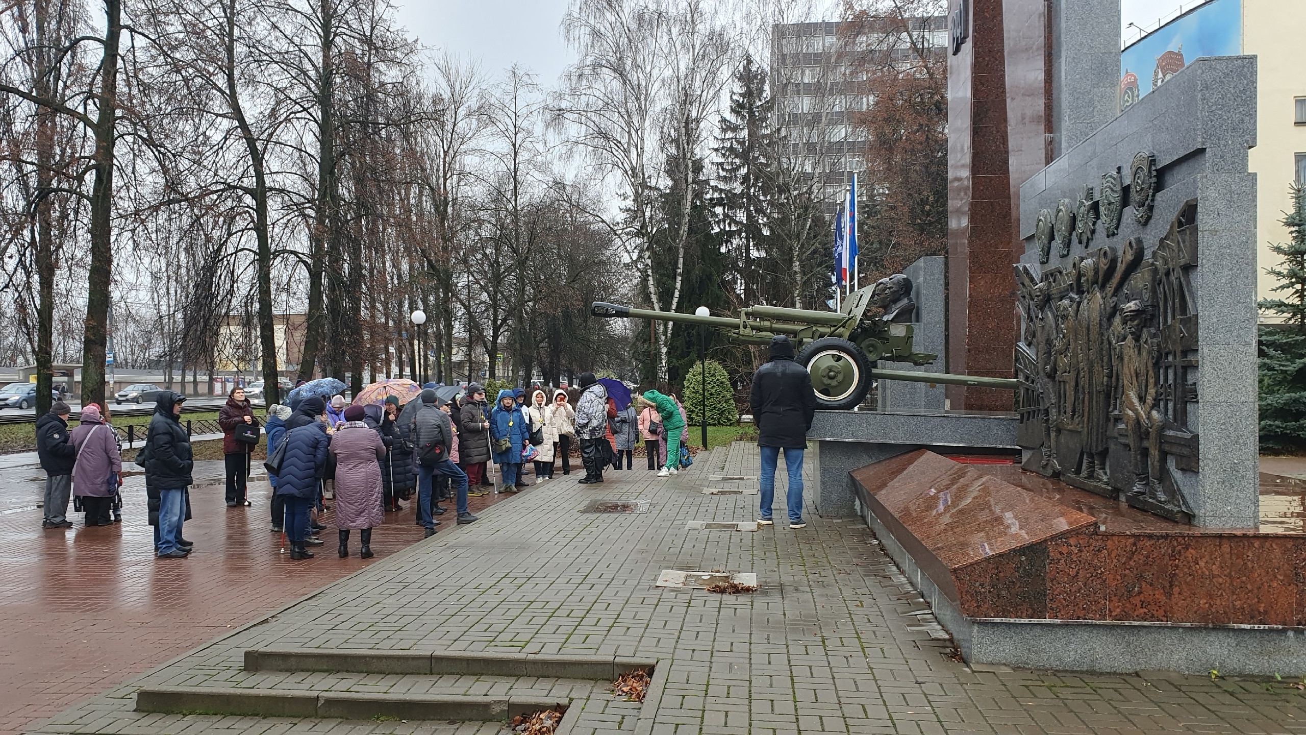 Памятник на городской площади. На переднем плане — артиллерийское орудие, установленное на постамент. Справа от пушки размещён крупный барельеф с изображением людей, занятых работой или в военной обстановке. Слева собралось около тридцати человек.