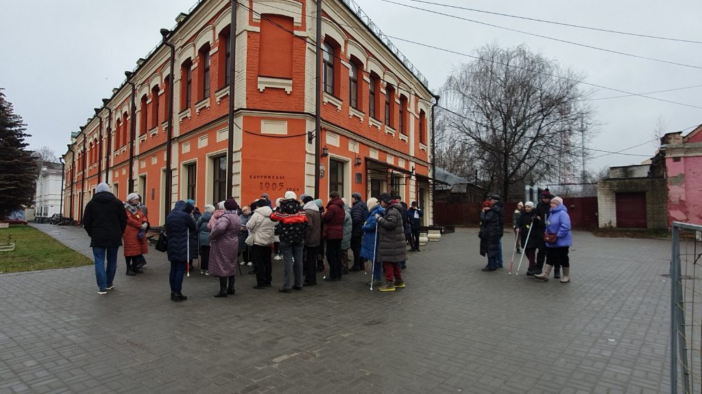 На фотографии изображено красно-белое двухэтажное здание с арочными окнами и декоративными элементами по фасаду. Угловая часть здания выступает вперёд.
На стене написано: "Баррикады 1905 года". Перед зданием собралось около двадцати человек, большинство одеты в тёплую зимнюю одежду — куртки, шапки, шарфы.
Несколько человек держат белые трости, предназначенные для людей с нарушением зрения.