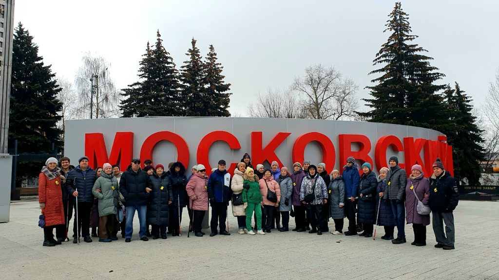 Группа людей стоит на улице перед большими красными буквами, образующими слово "МОСКОВСКИЙ" на сером фоне. Почти все одеты в зимнюю одежду — куртки, пальто,
шапки и шарфы, некоторые держат трости. Позади них видны высокие ели, голые деревья и пасмурное небо. Некоторые люди улыбаются, а сама группа выглядит
организованной для общего фото, возможно, на экскурсии или мероприятии.