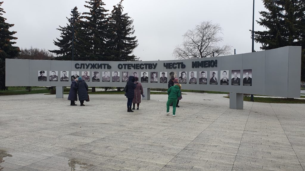 Мемориальная стена с черно-белыми портретами мужчин, большинство из которых в военной форме. В верхней части крупная надпись: "СЛУЖИТЬ ОТЕЧЕСТВУ ЧЕСТЬ
ИМЕЮ!" На площади перед стеной находятся несколько человек: взрослые и дети, одетые в зимнюю одежду. Фон украшен хвойными деревьями и облачным небом, атмосфера
пасмурная. Мемориал посвящён людям, служившим своему Отечеству.