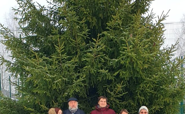 На фотографии изображены пятеро взрослых людей, стоящих на снегу перед огромной лесной елью. Ёлка занимает почти весь фон — она густая, пушистая, зелёная, выше нескольких человеческих ростов, ветви раскинуты широко в стороны и вверх, местами видны свисающие шишки. Вокруг ровный слой чистого белого снега. Небо светлое и затянуто тучами.