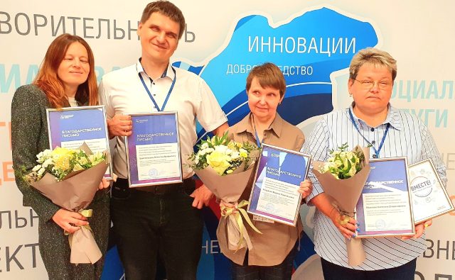 Сотрудники Центра "Камерата" стоят на фоне стенда с цветами и благодарственными письмами.
