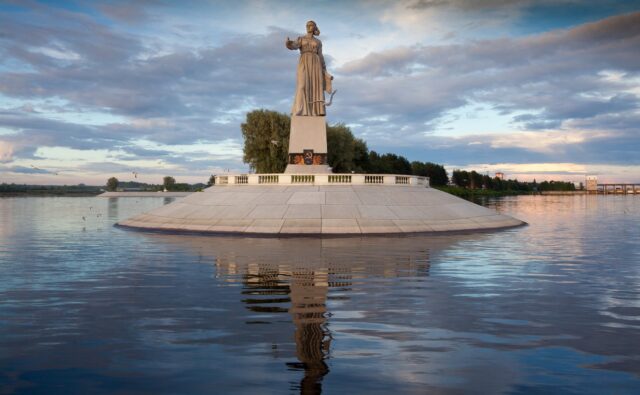 На фотографии изображен памятник "Мать-Волга" в Рыбинске, Россия. Памятник стоит на круглом постаменте, который окружен водой. Постамент выполнен из светлого камня и имеет наклонные грани, ведущие к основанию. В центре постамента возвышается фигура женщины в длинном платье, символизирующая Волгу. Женщина стоит с поднятой правой рукой, в которой держит голубя, символ мира. Левой рукой она придерживает край платья. На передней части постамента виден барельеф с изображением серпа и молота, символа Советского Союза, окруженного лучами. Вокруг постамента установлено ограждение в виде невысокого парапета с балюстрадой. Вода вокруг постамента спокойная, и в ней четко отражается фигура памятника и небо. На заднем плане видны деревья и кустарники, создающие зеленый фон. Небо покрыто облаками, которые отражаются в воде, создавая живописный вид. В небе и над водой летают несколько чаек, добавляя динамики в сцену. На дальнем плане, справа, видны элементы городской инфраструктуры, такие как здания и мостовые конструкции, что указывает на близость города. В целом, фотография передает спокойствие и величие памятника, гармонично вписанного в природный и городской ландшафт.