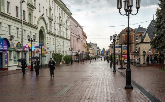 На фотографии изображена пешеходная улица в городе. Слева и справа расположены здания с архитектурными элементами, характерными для старинных построек. На улице установлены фонари в ретро-стиле. Люди идут по улице, некоторые из них держат зонты, что указывает на дождливую погоду. На переднем плане слева видны вывески магазинов и аптек. Вдали виднеется зелёная крыша здания, возможно, это часть исторического центра города.