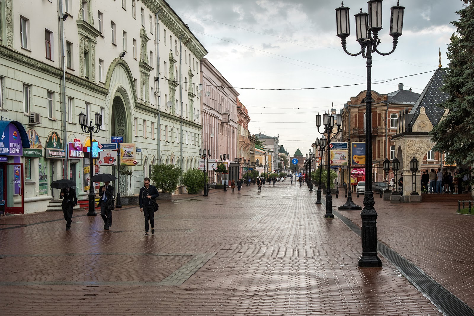 На фотографии изображена пешеходная улица в городе. Слева и справа расположены здания с архитектурными элементами, характерными для старинных построек. На улице установлены фонари в ретро-стиле. Люди идут по улице, некоторые из них держат зонты, что указывает на дождливую погоду. На переднем плане слева видны вывески магазинов и аптек. Вдали виднеется зелёная крыша здания, возможно, это часть исторического центра города.