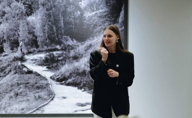 Женщина в чёрном пиджаке на фоне чёрно-белой фотографии. Она стоит в помещении и что-то рассказывает или объясняет, жестко выражая эмоции рукой. На фоне — большая чёрно-белая фотография с изображением извилистой тропинки среди деревьев, скорее всего, в галерее или музее.