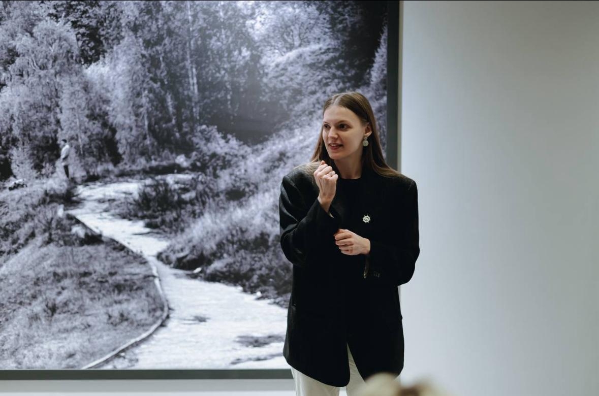 Женщина в чёрном пиджаке на фоне чёрно-белой фотографии. Она стоит в помещении и что-то рассказывает или объясняет, жестко выражая эмоции рукой. На фоне — большая чёрно-белая фотография с изображением извилистой тропинки среди деревьев, скорее всего, в галерее или музее.