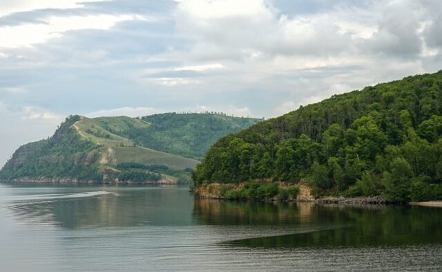 На фотографии изображен спокойный природный пейзаж. В центре кадра находится водоем, возможно, река или озеро, с гладкой поверхностью воды, которая отражает небо и окружающие деревья. Вода темная, почти черная, с легкими рябями от движения лодки или катера, который оставил за собой след в виде белых волн, уходящих влево. На переднем плане, справа, виден берег, покрытый густым лесом. Деревья высокие, с пышной зеленой листвой, создающей плотную стену зелени. Берег обрывистый, с видимыми участками земли и камней, которые местами оголены. На заднем плане, слева, возвышается холм, покрытый травой и редкими деревьями. Холм имеет плавные очертания и поднимается вверх, создавая ощущение глубины и перспективы. На его склонах видны светлые участки, возможно, это тропинки или открытые участки земли. Небо покрыто облаками, которые создают мягкое, рассеянное освещение. Облака светло-серые, местами белые, что придает небу объем и текстуру. В целом, фотография передает ощущение спокойствия и умиротворения, характерное для природных ландшафтов.