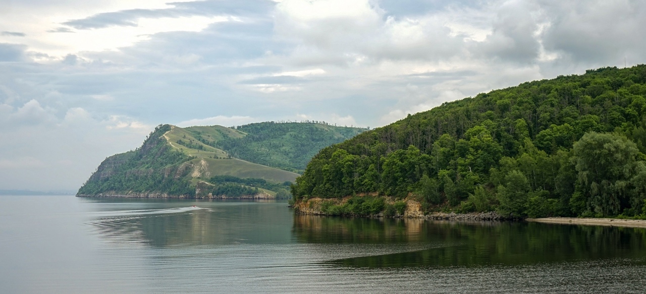 На фотографии изображен спокойный природный пейзаж. В центре кадра находится водоем, возможно, река или озеро, с гладкой поверхностью воды, которая отражает небо и окружающие деревья. Вода темная, почти черная, с легкими рябями от движения лодки или катера, который оставил за собой след в виде белых волн, уходящих влево. На переднем плане, справа, виден берег, покрытый густым лесом. Деревья высокие, с пышной зеленой листвой, создающей плотную стену зелени. Берег обрывистый, с видимыми участками земли и камней, которые местами оголены. На заднем плане, слева, возвышается холм, покрытый травой и редкими деревьями. Холм имеет плавные очертания и поднимается вверх, создавая ощущение глубины и перспективы. На его склонах видны светлые участки, возможно, это тропинки или открытые участки земли. Небо покрыто облаками, которые создают мягкое, рассеянное освещение. Облака светло-серые, местами белые, что придает небу объем и текстуру. В целом, фотография передает ощущение спокойствия и умиротворения, характерное для природных ландшафтов.