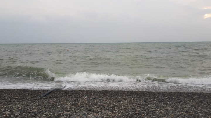 На фотографии изображен морской берег. Вода слегка волнуется, волны накатывают на гальку, покрывающую пляж. Небо облачное, серое, без яркого солнца. Вдалеке на горизонте видна линия, где море встречается с небом.